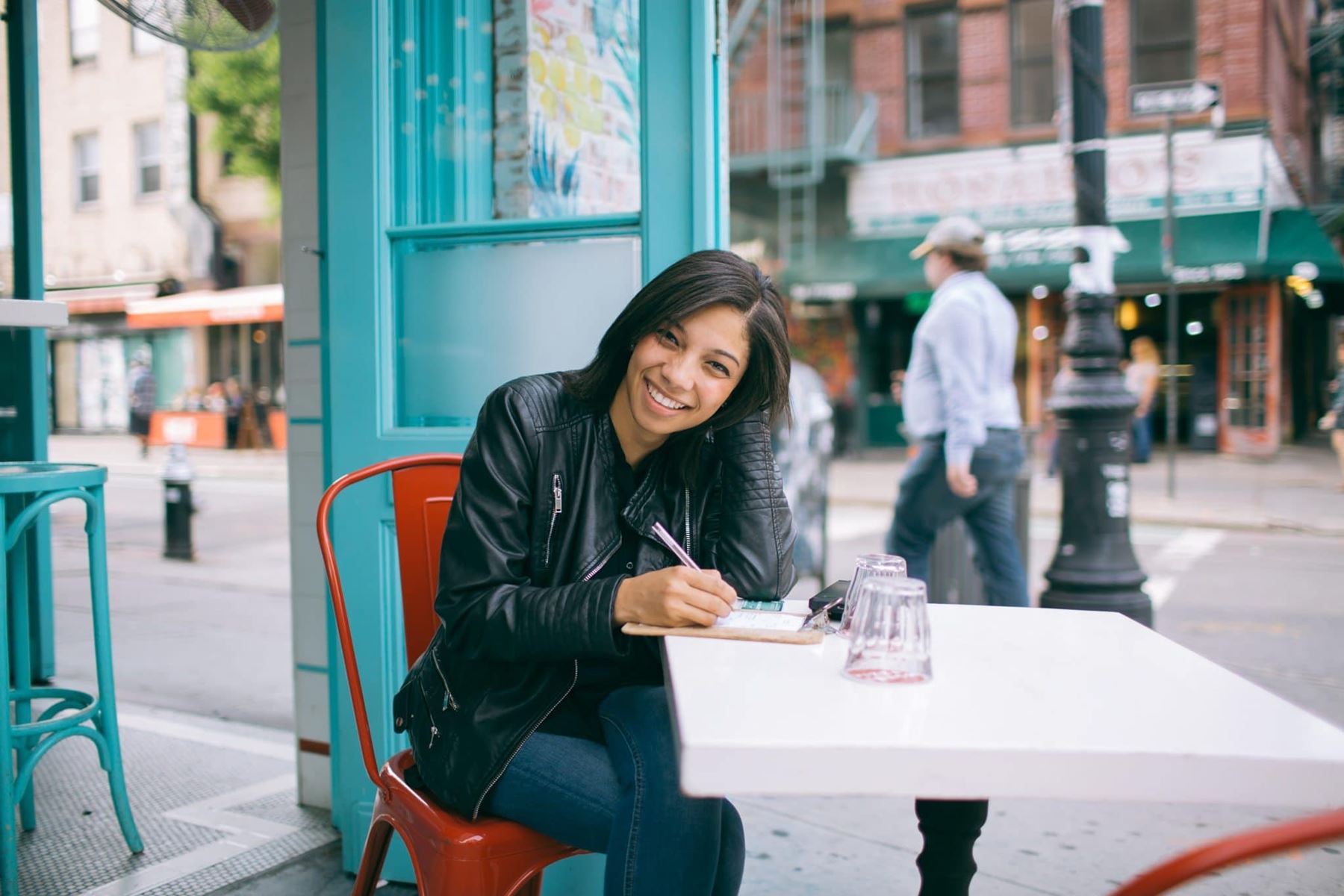 Woman sitting at a table outside smiling towards the camera and writing on a notepad